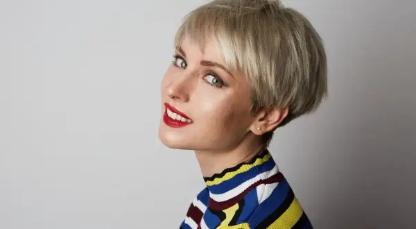 Kobieta z krótkimi blond włosami w stylu pixie, w kolorowym golfie, z czerwonymi ustami.